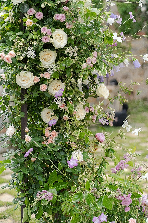 Floral wedding arch with white roses and greenery vines, accented by lavender blooms, set on a garden lawn near trees and stone building