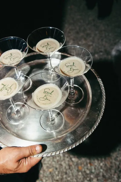 Signature cocktails in martini glasses with personalized cocktail foam and printed toppers, served on a silver tray over stone pavement