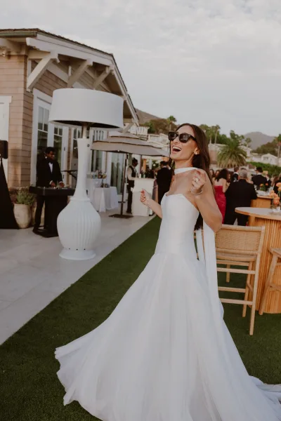 Bride portrait in sunglasses and a bridal gown on a palm-lined lawn with guests by a bar and distant mountain views.