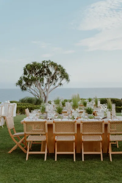 Reception tablescape featuring rattan chairs and elegant outdoor reception decor on a lawn, set beside an open ocean horizon under clear sky.