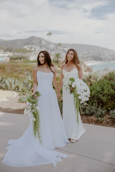 Couple portrait of brides holding hands on a coastal pathway, two brides in white with orchid bouquets, ocean and green hills stretching behind.