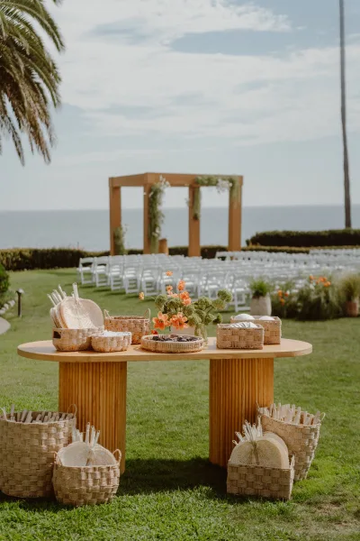 Wedding welcome table serving as a ceremony welcome station, with woven baskets, set beside the ocean with a palm tree, arch, and white chairs.