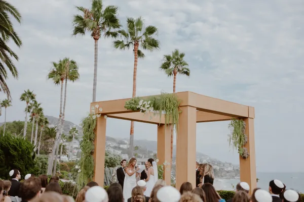 Wedding vows shared in an outdoor ceremony moment beneath a wooden canopy, with palm trees and ocean beyond