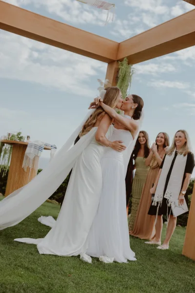 Ceremony kiss between two brides at a same-sex wedding ceremony beneath a chuppah, captured in soft natural light.