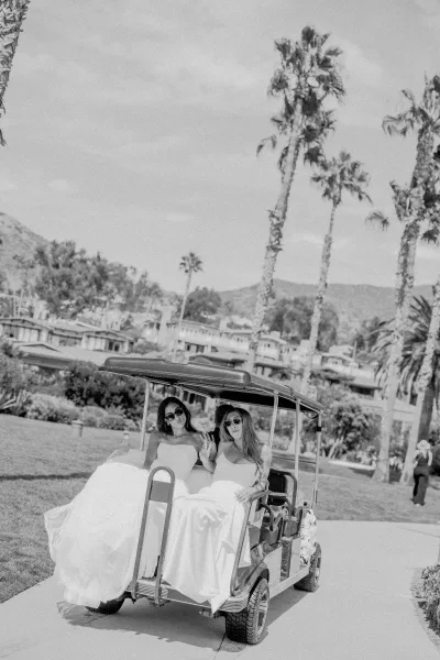 Golf cart wedding photo capturing a candid wedding moment as the bridal party rides past palm trees, the bride in sunglasses, mountains beyond.