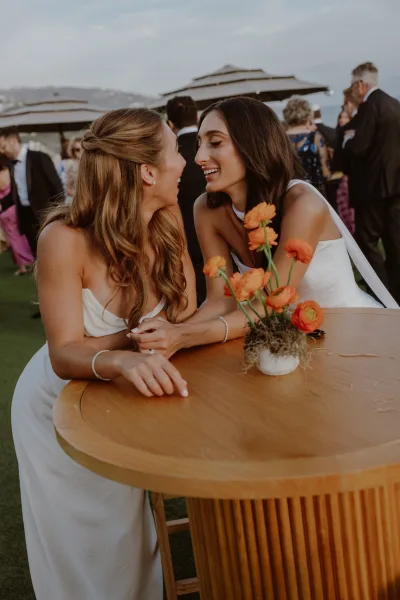 Cocktail hour moment with brides laughing together as two brides lean on a cocktail table with an orange ranunculus bud vase beneath patio umbrellas.