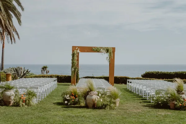 Ceremony setup featuring outdoor ceremony decor with a wooden square arch on a grass lawn, facing the ocean under an open sky.
