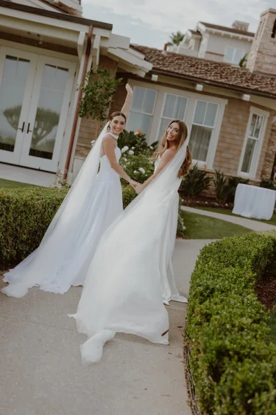 Couple portrait, a two brides wedding photo of them holding hands along a garden walkway, matching veils flowing behind as they walk together.