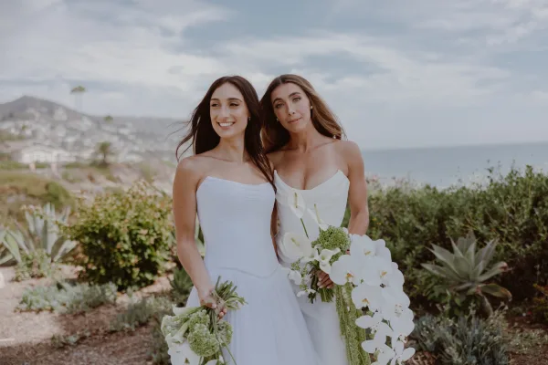 Couple portrait of two brides on a coastal bluff above the ocean, holding a white orchid bouquet beside sunlit succulents.