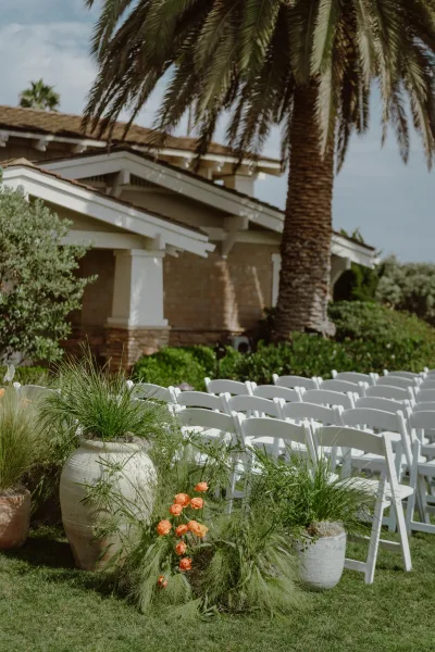 Ceremony aisle decor for outdoor ceremony seating, accented with potted greenery along the aisle, set on a sunny lawn beneath a palm.