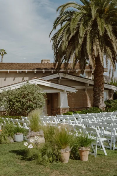 Outdoor ceremony setup with a refined ceremony seating layout of white folding chairs on a green lawn, backed by palm trees and a house exterior.