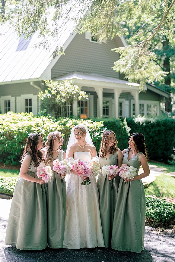 Bridal party portrait of the bride with bridesmaids in sage green dresses, holding bouquets on a garden walkway with greenery and a house behind