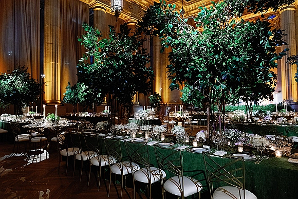 Reception tablescape with green wedding tablescape linens, tall greenery centerpieces, and candles set in a grand hall with columns and uplighting