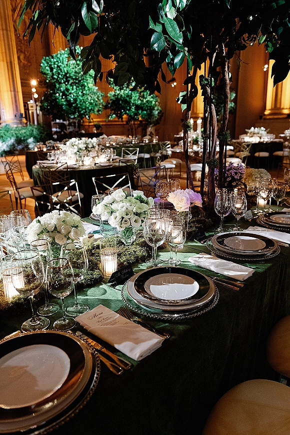 Reception tablescape with green wedding table decor, white rose and tree centerpieces, moss runner, votive candles, and gold flatware in a warm ballroom
