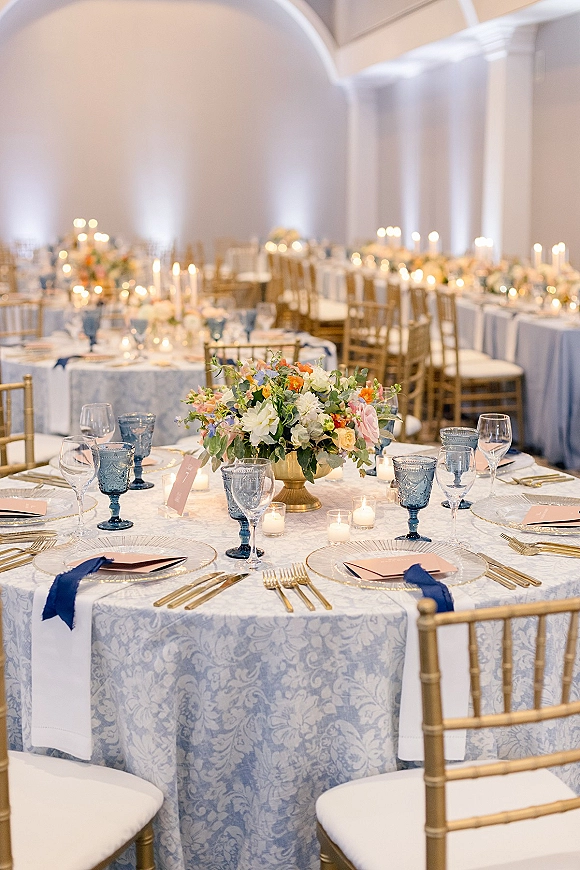 Reception tablescape with a wedding table centerpiece in a gold compote vase, blue goblets, candles, and place cards in a softly lit banquet hall