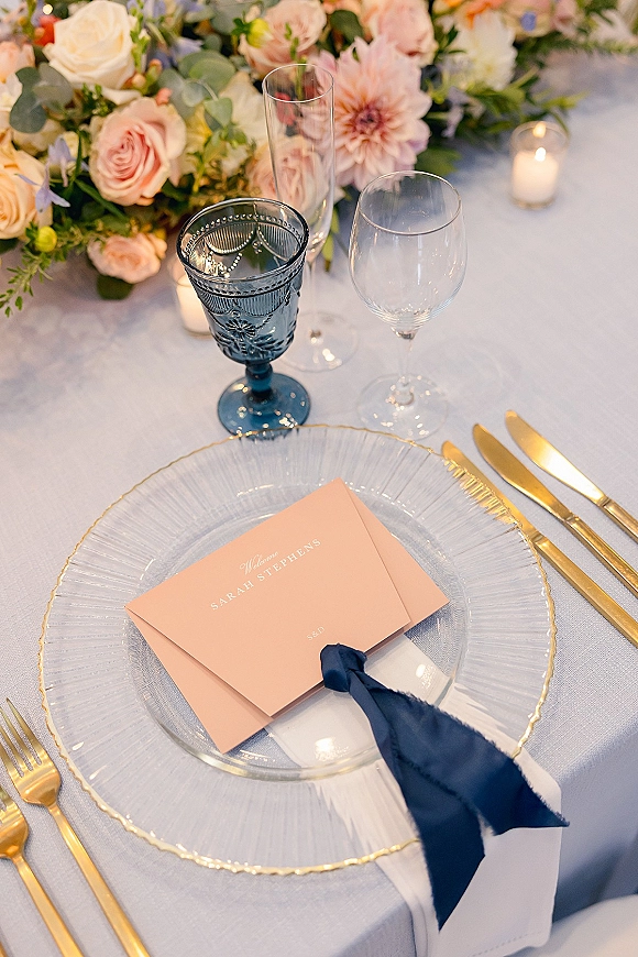 Reception place setting with wedding place card on a glass charger plate, navy napkin, gold flatware, blue goblet, candles and floral centerpiece