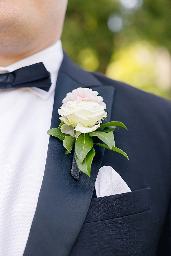 Groom boutonniere with a white rose and pink accent pinned to a black tuxedo lapel, with greenery leaves against blurred outdoor foliage
