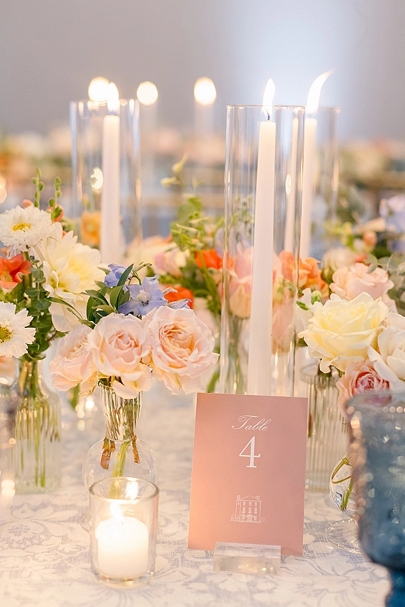 Reception tablescape with wedding table centerpiece of taper candles in glass, pastel roses and greenery garland on lace tablecloth, warm bokeh background