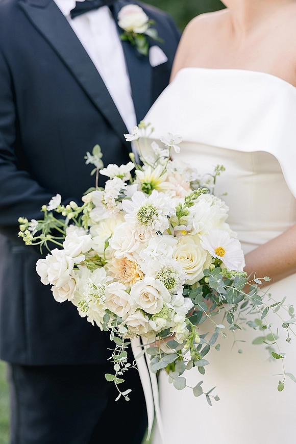 Wedding bouquet of white and ivory blooms with roses, daisies, hydrangea and eucalyptus, held by bride beside groom in tuxedo outdoors