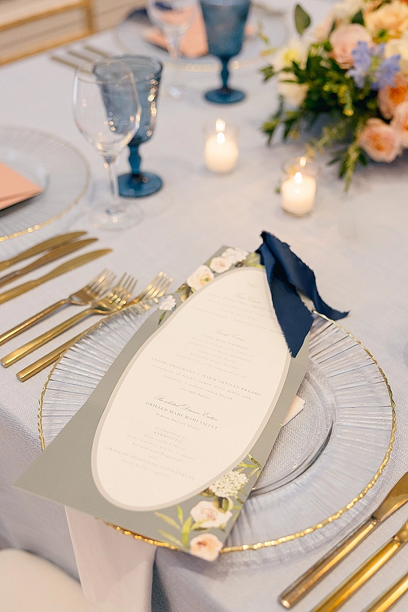 Reception tablescape with a wedding place setting, menu card tied with navy ribbon on white plates, gold flatware, blue goblets, candles and florals