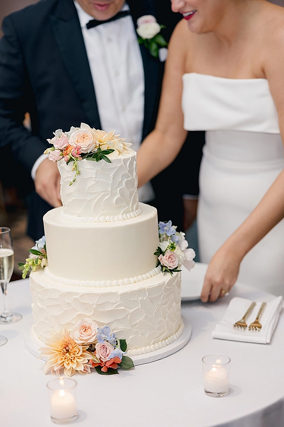 Wedding cake cutting as bride in strapless gown and groom in tux slice a three-tier cake with floral topper at candlelit reception table