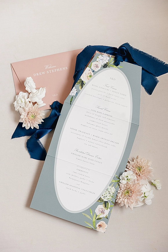 Wedding menu card with floral illustrations tied in a navy ribbon, styled with blush and white flowers and a classical bust on a neutral backdrop