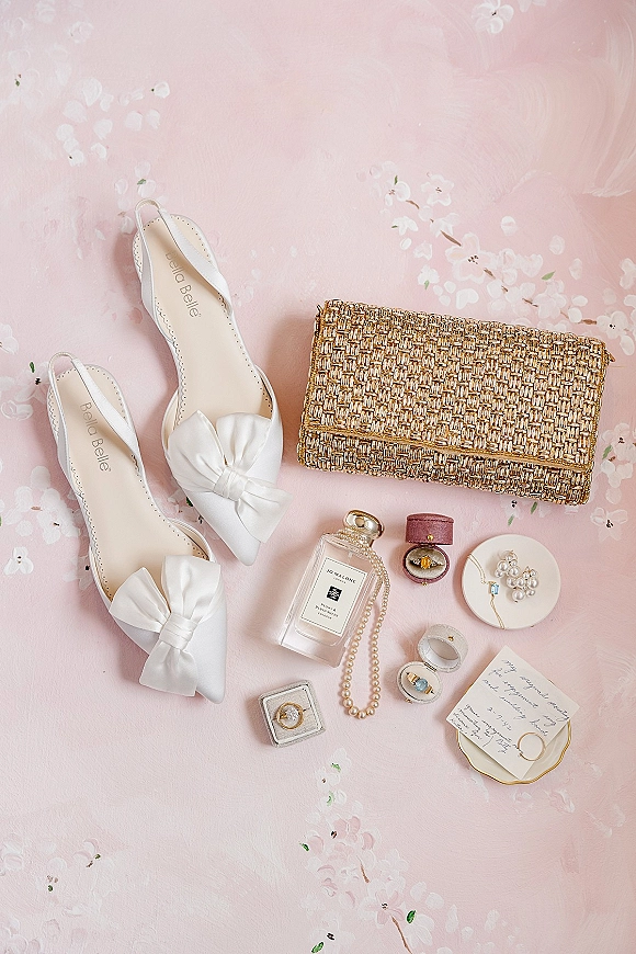 Bridal accessories flat lay with wedding details flat lay: white bow slingbacks, gold beaded clutch, perfume, pearls, rings on pink floral surface