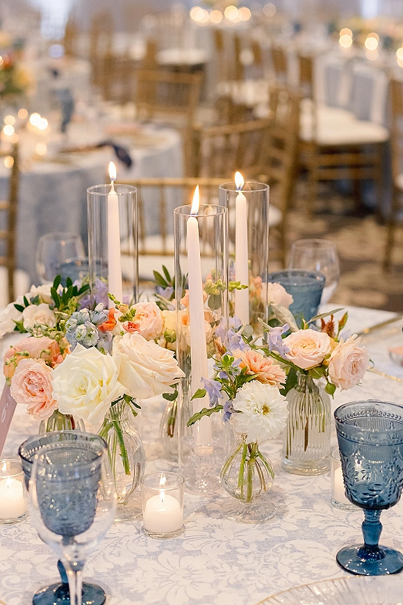 Reception tablescape with wedding table centerpiece of pastel flowers, white taper candles in glass holders, blue goblets, and bistro lights overhead