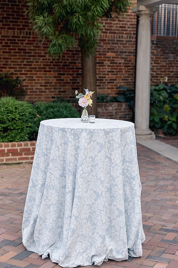 Cocktail table decor with pastel bud vase flowers, greenery sprig, and votive candle on patterned cloth in a brick courtyard setting
