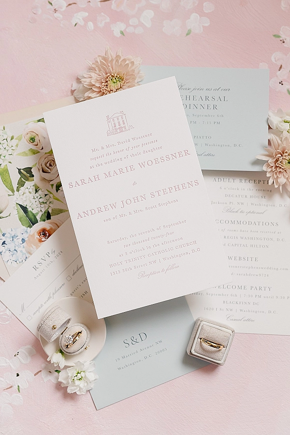 Wedding invitation suite with blush wedding invitations, RSVP card, floral envelope liner, ring boxes and rings arranged on a pink surface
