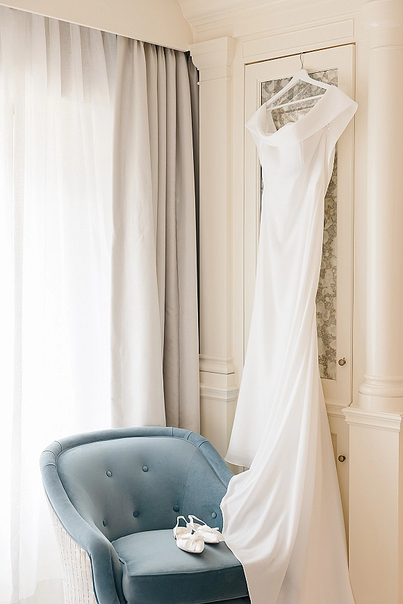 Wedding dress hanging by a window, a white bridal gown on hanger beside bridal shoes on a blue velvet chair in soft light