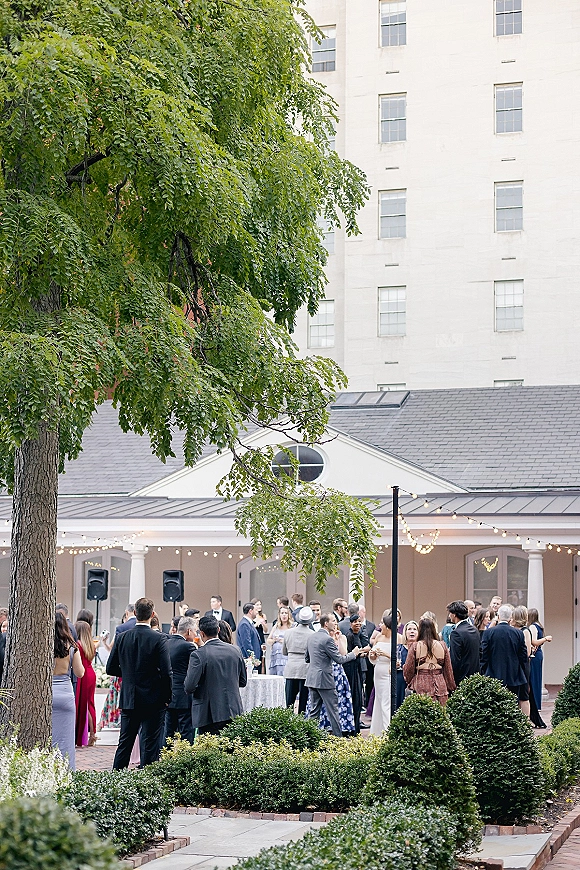 Wedding cocktail hour with guests mingling around cocktail tables with linens under string lights in a brick courtyard with a large tree