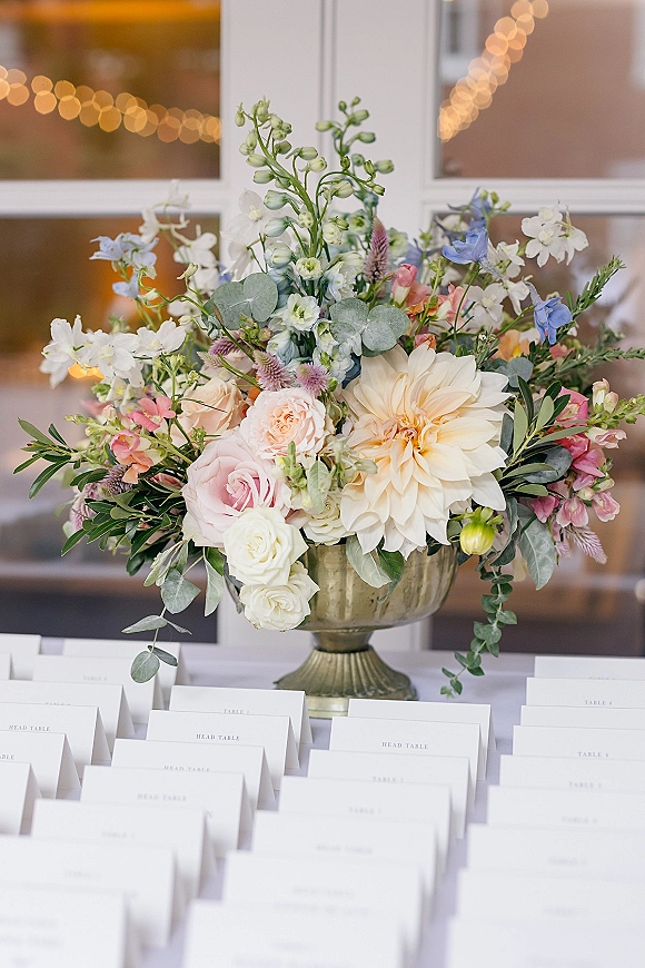 Escort card display with wedding escort cards arranged beside a compote vase of roses, dahlia, and eucalyptus under string lights by windows