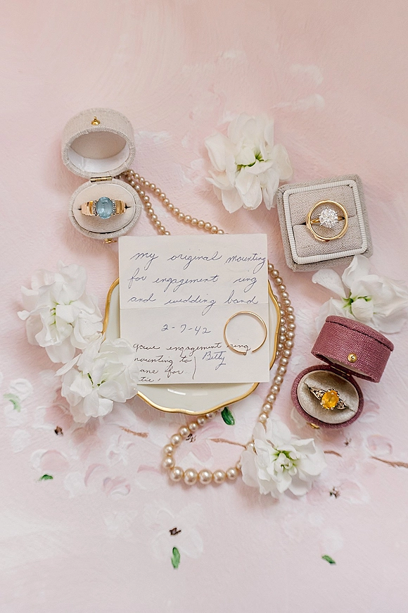 Wedding rings flatlay with engagement ring and band in ring boxes, pearl necklace and handwritten note on pink fabric with petals