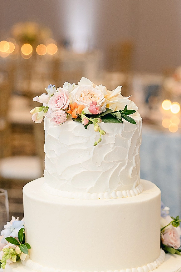 Wedding cake with two tier wedding cake design, textured white buttercream and fresh roses and peony topper in a bokeh-lit reception room