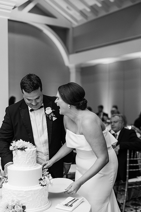 Wedding cake cutting as bride in strapless dress and groom in tuxedo slice a tiered floral cake at indoor reception with guests behind