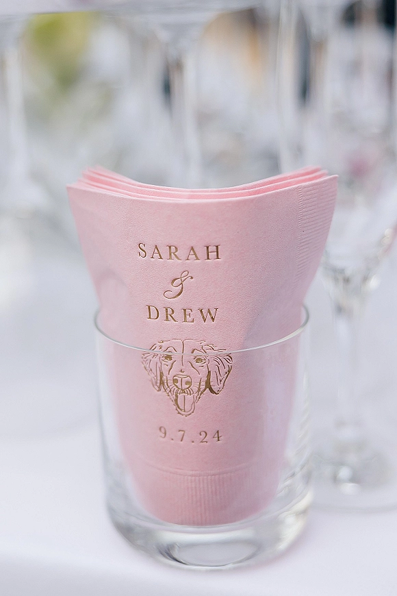 Wedding cocktail napkins with personalized cocktail napkins in pink paper, gold foil lettering and dog illustration beside a clear glass on white tablecloth