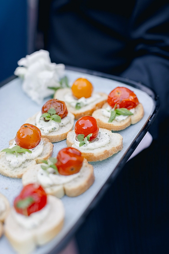 Wedding cocktail appetizers on a tray with crostini topped with herbed cheese, cherry tomatoes, microgreens, and a white rose accent