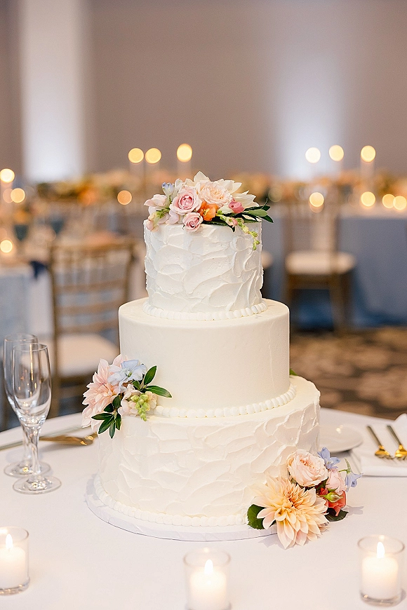 Wedding cake with three tier design, white buttercream frosting and blush, peach, and light blue florals on a candlelit reception table