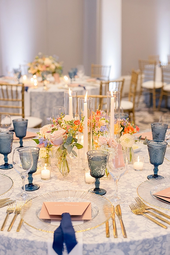 Reception tablescape with wedding table centerpiece of pastel florals, taper candles, blue goblets and gold flatware on patterned cloth in uplit room