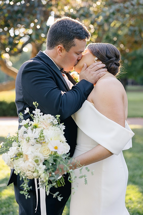 Wedding kiss portrait of bride and groom kissing, her white off-the-shoulder dress and bouquet, in a sunlit garden with bokeh background