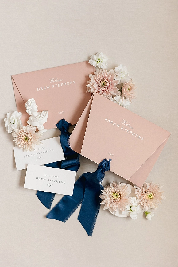 Invitation suite with blush wedding invitations, dusty pink envelopes, navy ribbon, place cards, flowers, and a ceramic bust on neutral surface