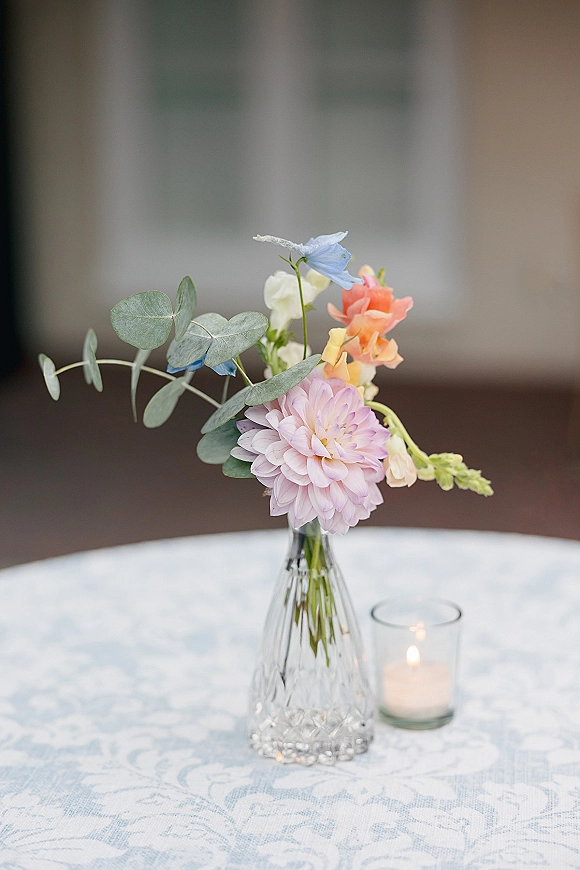 Bud vase centerpiece with a pastel flower and eucalyptus greenery beside a votive candle on a white patterned tablecloth, blurred facade behind