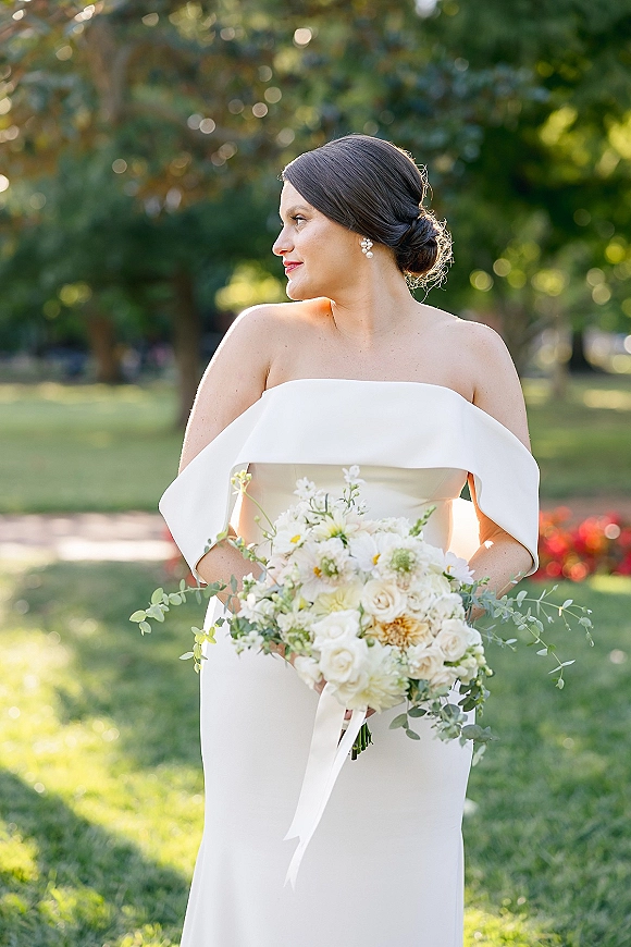 Bridal portrait of a bride in an off the shoulder wedding dress holding a white rose and daisy bouquet in a sunlit garden with trees