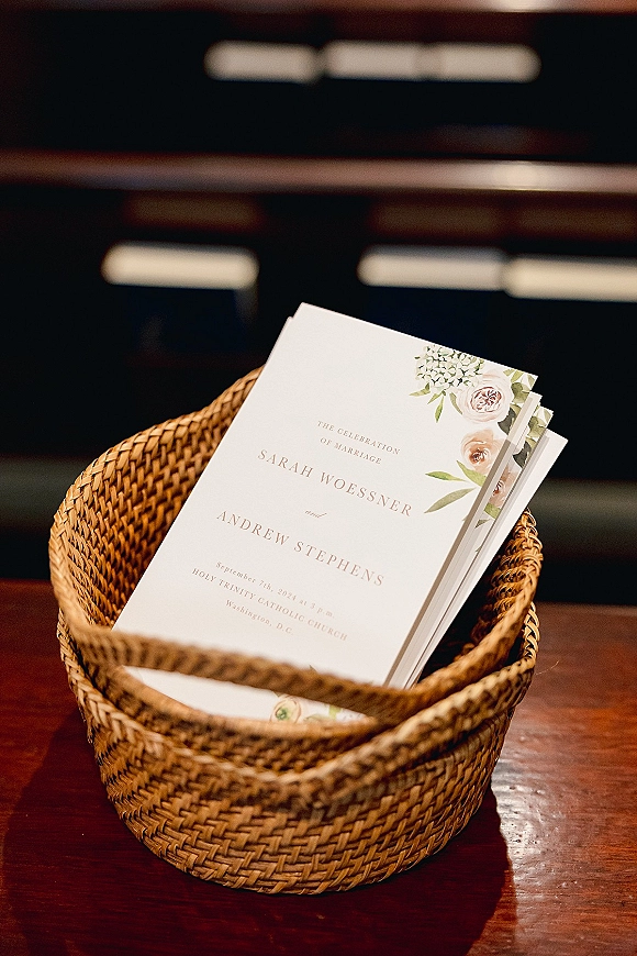 Wedding programs stacked in a woven basket, wedding ceremony program cards with floral illustration on a wood table near church pews