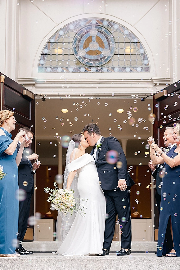 Wedding kiss portrait with bubble send off as bride in veil holds bouquet and kisses groom in tuxedo at church entrance under stained glass
