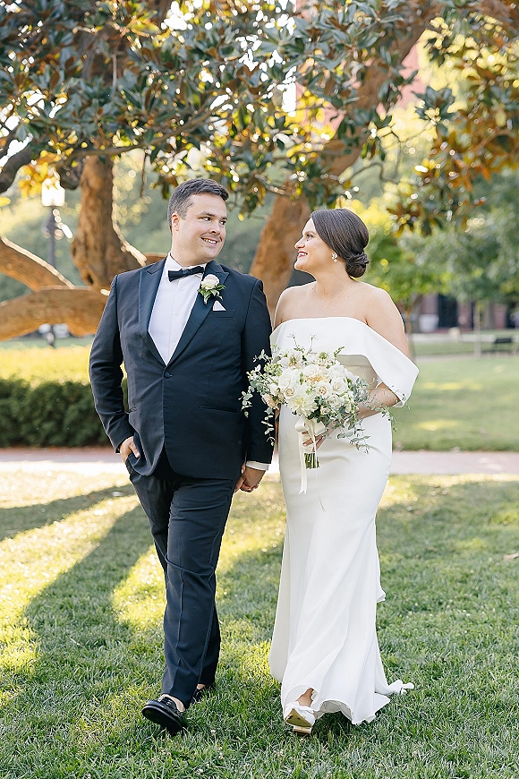 Couple portrait of bride and groom walking hand in hand on a sunny park path, her bouquet ribbon trailing beside his black tuxedo