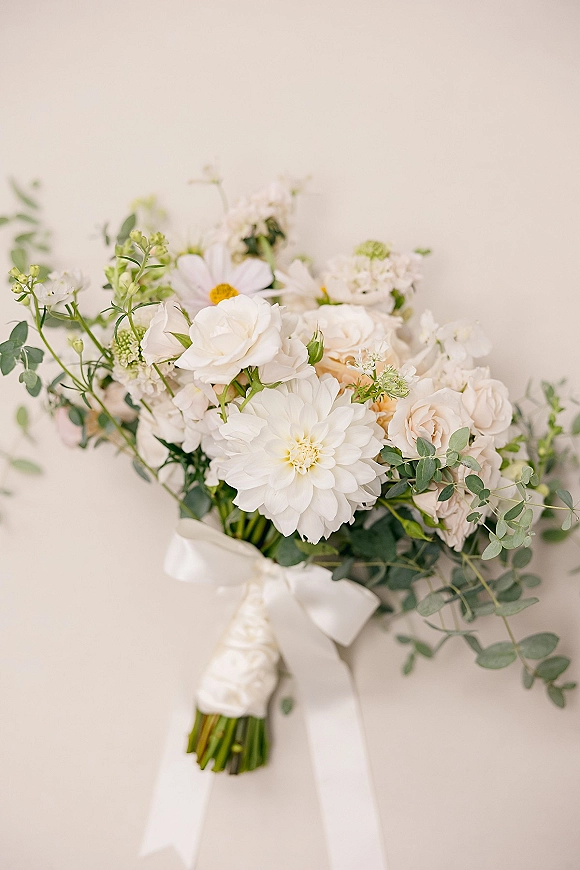 Bridal bouquet of white and blush bouquet blooms with dahlia, daisies, and eucalyptus, finished with a white satin ribbon against a neutral wall