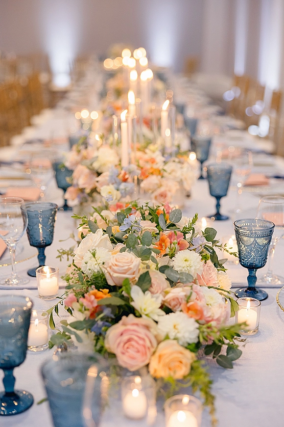 Reception tablescape with wedding table centerpiece featuring a floral table runner, taper and votive candles, and blue goblets in a draped, uplit room