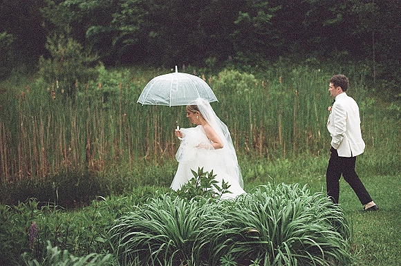 Wedding couple portrait with bride holding a clear umbrella, veil flowing as groom in tuxedo follows through lush garden greenery in rain